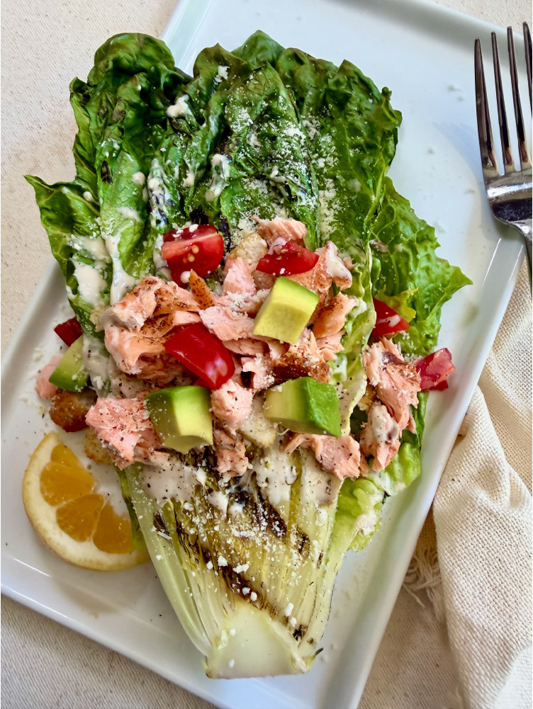 Close-up of Grilled Salmon Caesar Salad topped with California King Salmon, avocado cubes, tomatoes, Parmesan cheese, and Caesar dressing.